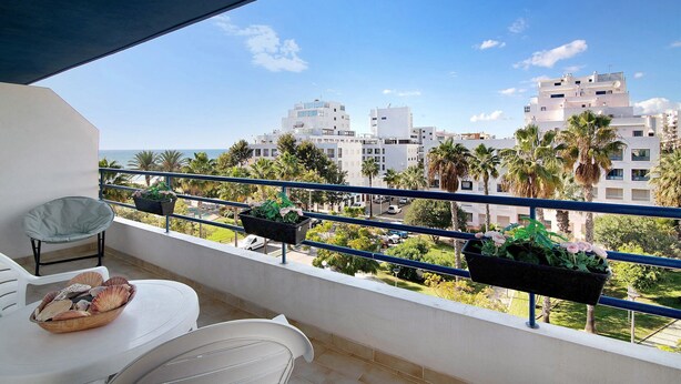 A balcony with a white chair, a small table with a bowl of seashells, and two flower planters.
