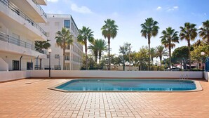 A swimming pool with a herringbone tile pattern, surrounded by palm trees and a multi-story building with balconies.