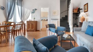 Een woonkamer met een bank, fauteuils, een eettafel met stoelen, een trap en een keukenhoek.