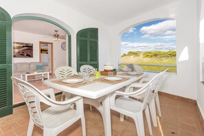 Un coin repas avec une table dressée pour quatre personnes, des chaises blanches, une vue par la fenêtre et une porte donnant sur une autre pièce.