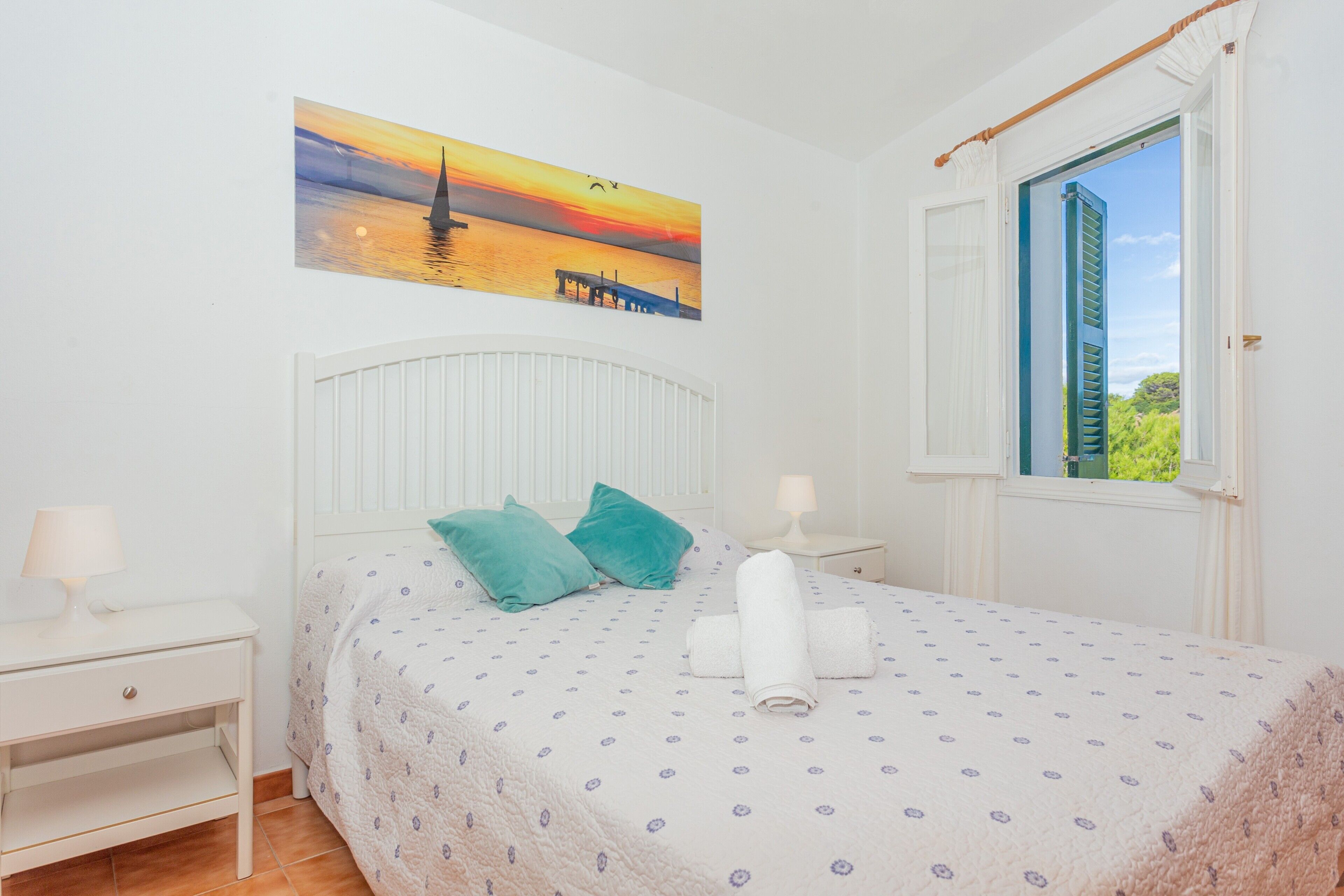 Une chambre à coucher avec un lit, des oreillers, une table de chevet, un tableau et une fenêtre avec des volets.