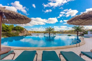 Ein Swimmingpool mit Liegestühlen und Strohdächern, umgeben von Grünpflanzen und blauem Himmel.