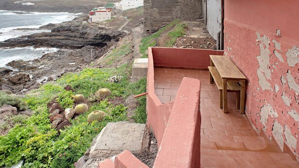 A coastal building with a red exterior, a wooden bench, and a view of the sea and rocky terrain.