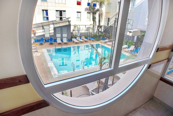 Vista de uma piscina com espreguiçadeiras e guarda-sóis, um deck revestido de azulejos e um edifício com janelas e varandas ao fundo.
