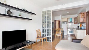 Uma sala de estar com televisão de tela plana, um sofá, uma área de refeições com cadeiras e uma cozinha com armários e eletrodomésticos.