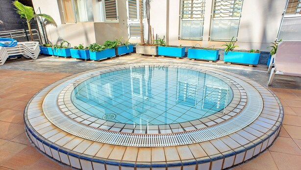 Uma piscina circular com borda azulejada, rodeada por vasos azuis com plantas e piso revestido de azulejos.
