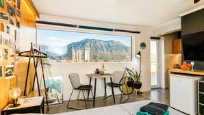 Una habitación moderna con un ventanal grande, una cama, un escritorio con una silla, una mesita y un refrigerador.