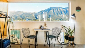 Una habitación con un ventanal grande, dos sillas, una mesa, una lámpara, una planta en maceta, un espejo y una maleta.