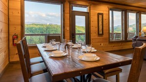 Una mesa de comedor de madera dispuesta con platos, vasos y cubiertos, con una vista panorámica de montañas a través de amplias ventanas.