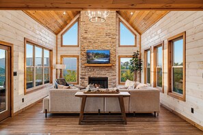 A spacious living room with a stone fireplace, large windows, and a high ceiling.
