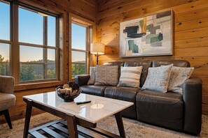 A cozy living room with a leather sofa, a coffee table, and a large window offering a view of the outdoors. - Panoramic Views, Hot Tub, Arcade Games, & Free Attraction Passes (Sevierville)