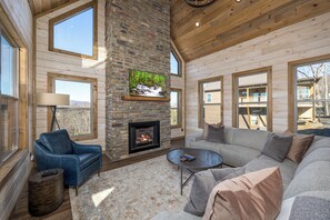 A cozy living room with a stone fireplace, a blue leather sofa, and a large window overlooking a deck.