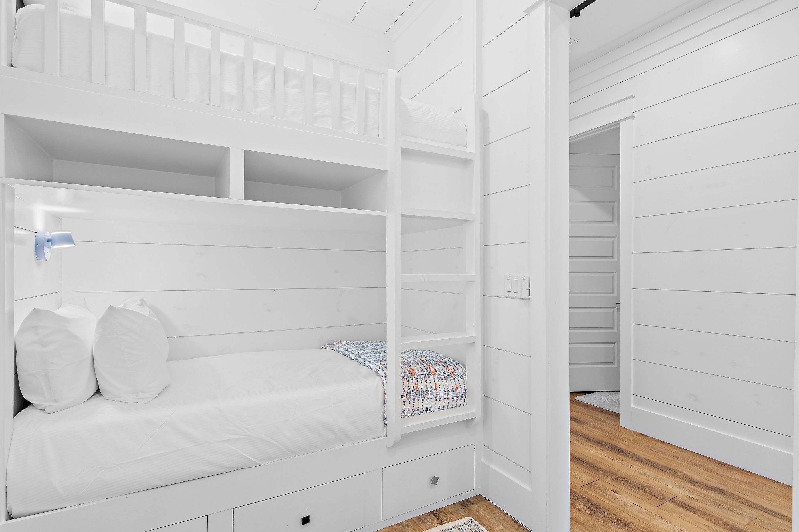 A white bunk bed with a patterned pillow, a small drawer, and a white door in the background.