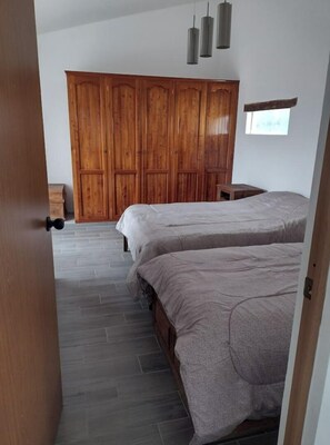 Two beds with grey bedding, a wardrobe, and a small table in a minimalist bedroom with a view.