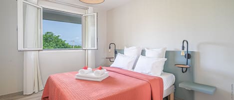 Una cama bien hecha con una colcha roja, una mesita de noche y una ventana con cortinas.
