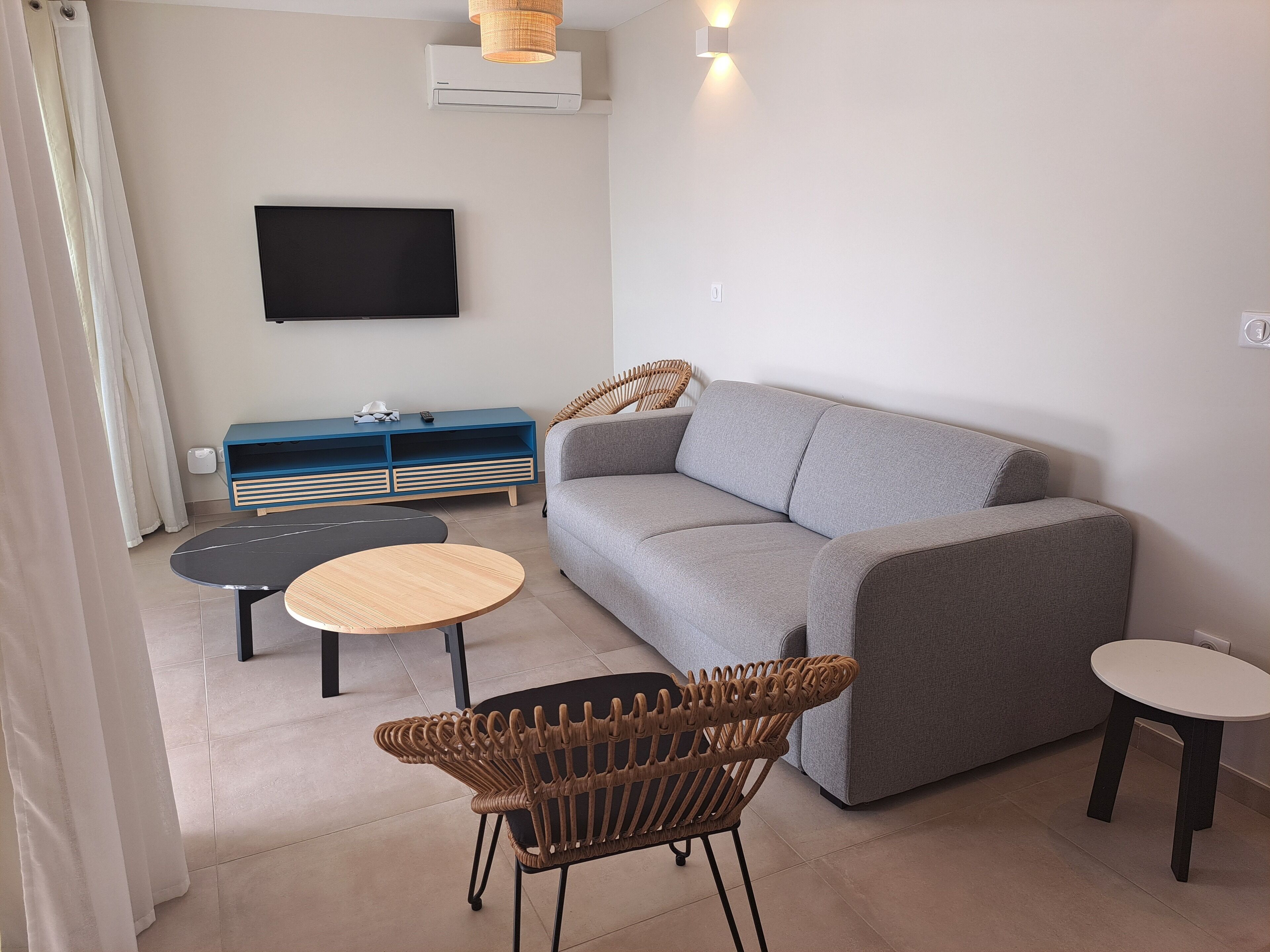 Ein modernes Wohnzimmer mit einem grauen Sofa, einer blauen Unterhaltungsanlage, einem Flachbildfernseher, einem Korbstuhl und zwei kleinen runden Tischen.