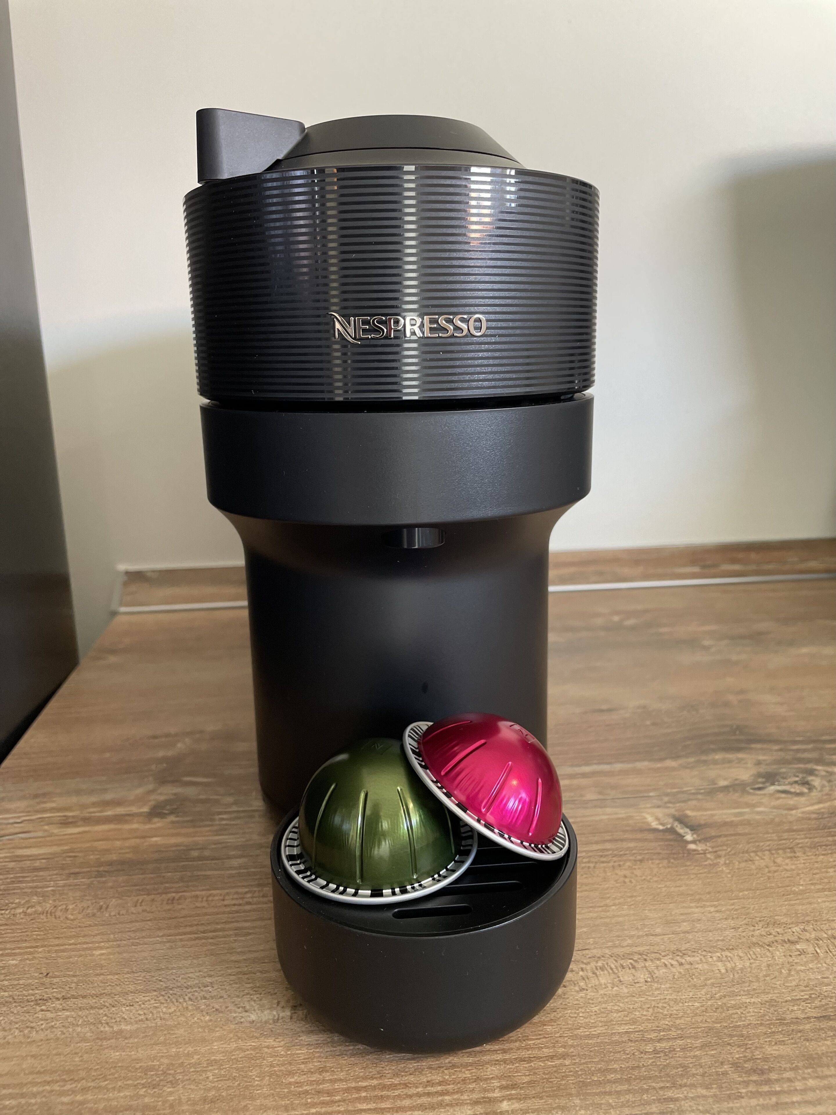 Une machine à café Nespresso noire, avec un couvercle et deux capsules, une verte et une rouge, posée sur une surface en bois.