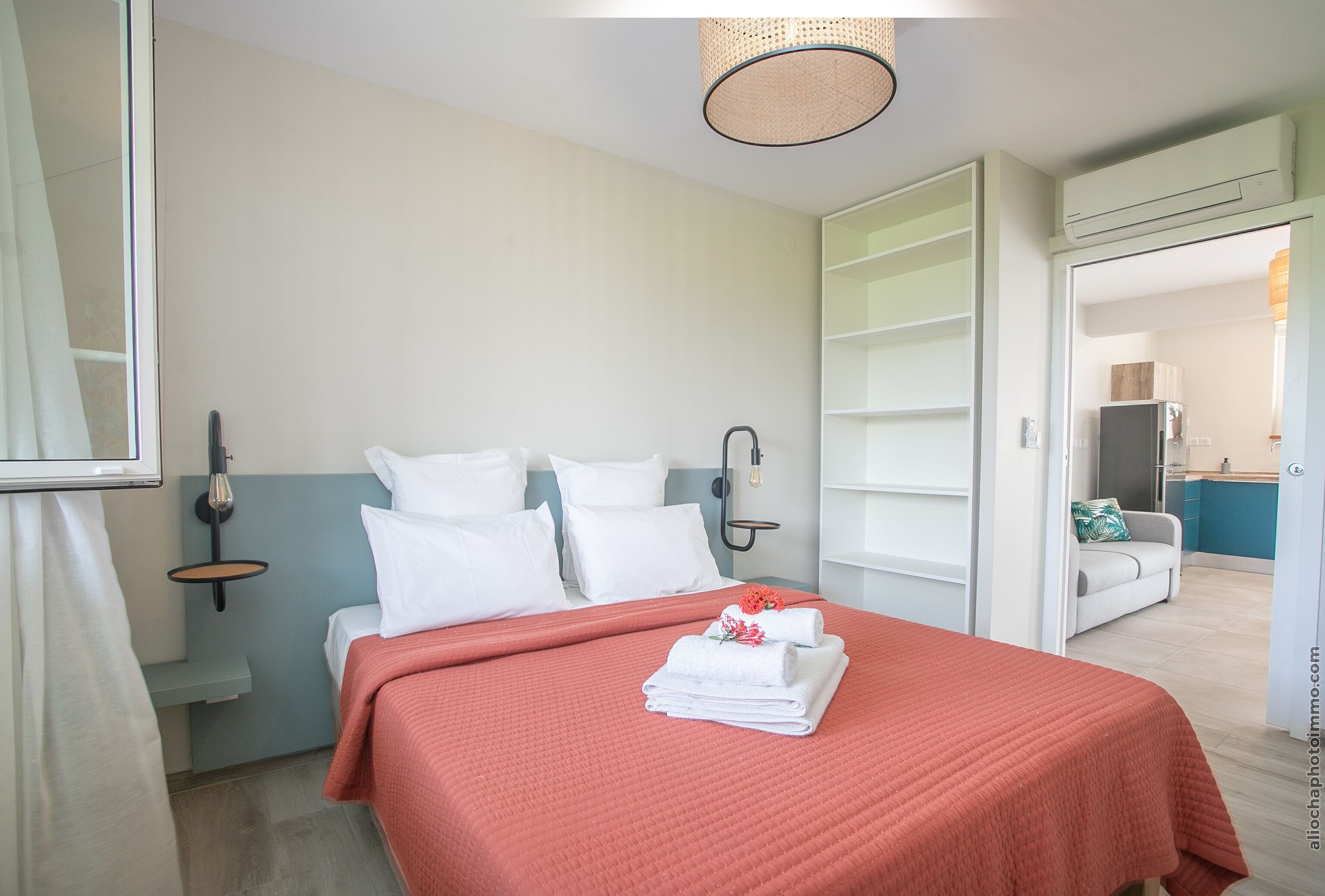 A hotel room with a bed, a nightstand, a wall-mounted lamp, and a view of the adjacent kitchen area.