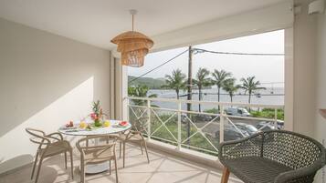 A balcony with a table set for two, a wicker chair, and a view of the sea and palm trees.