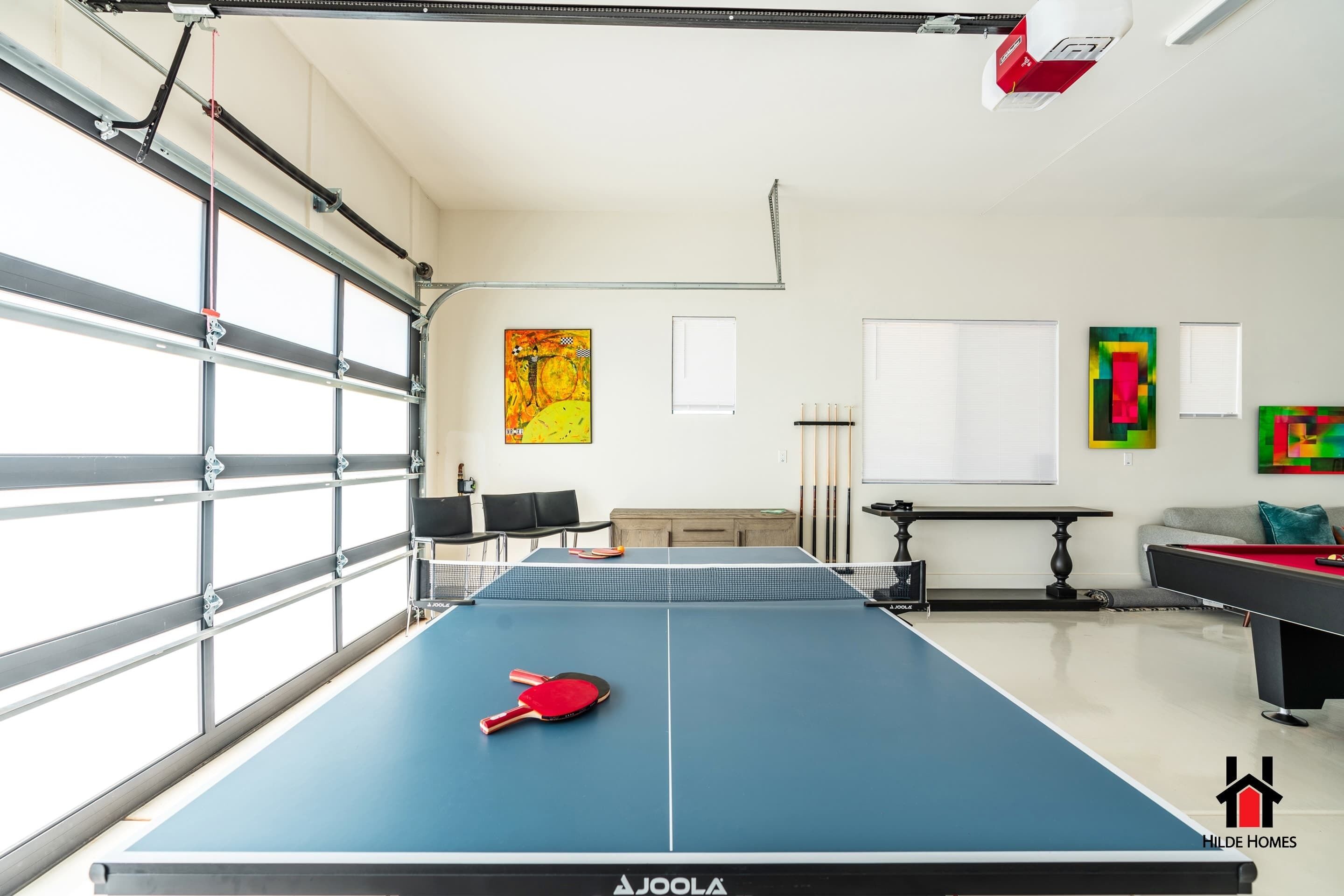 Une table de ping-pong avec raquettes, une table de billard et une table de jeu de fléchettes dans une pièce dotée de grandes fenêtres et ornée d’œuvres d’art.