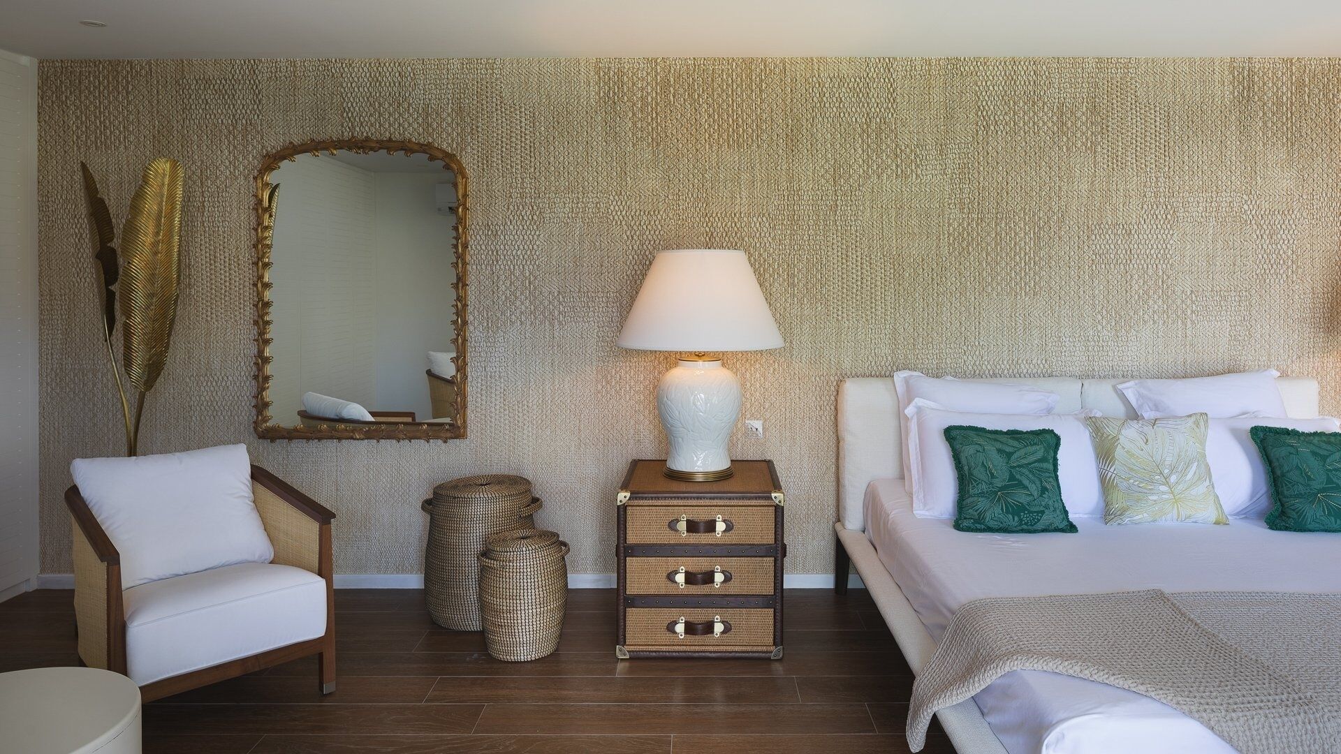Un dormitorio con un espejo grande, una lámpara de noche, una cama con almohadas blancas y verdes, una mesita de mimbre y una silla.