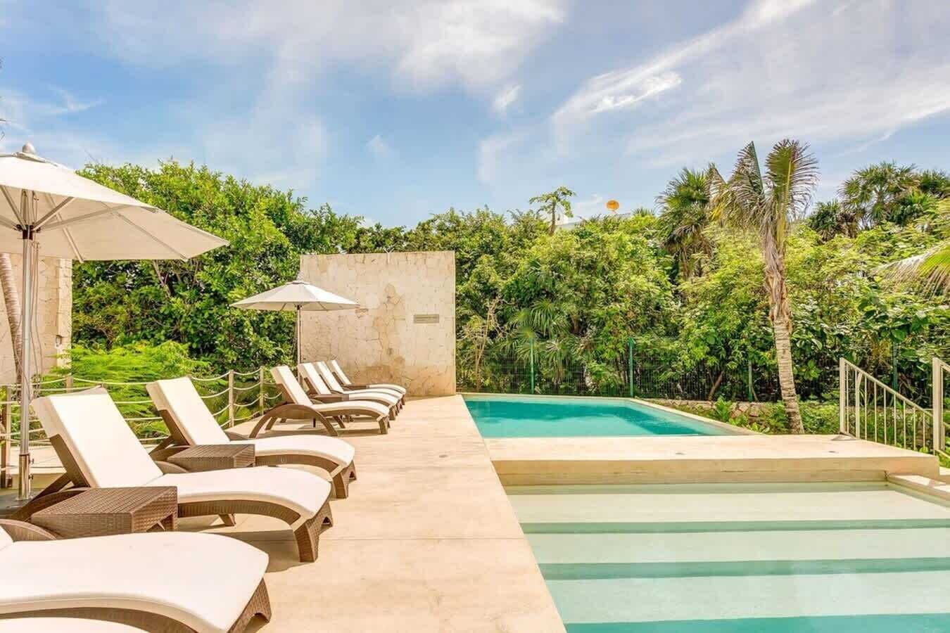 A poolside area with lounge chairs and umbrellas, surrounded by lush greenery.