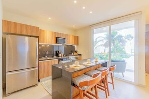 A modern kitchen with a central island, stainless steel appliances, and a view of palm trees through large sliding doors. - Ocean-View Patio, 9 Pools & Beach Club (Akumal)