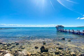 A clear blue sea with a rocky shore, a wooden pier, and a thatched-roof structure.