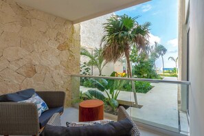 A balcony with wicker furniture, a wooden stool, and a view of palm trees and a building.