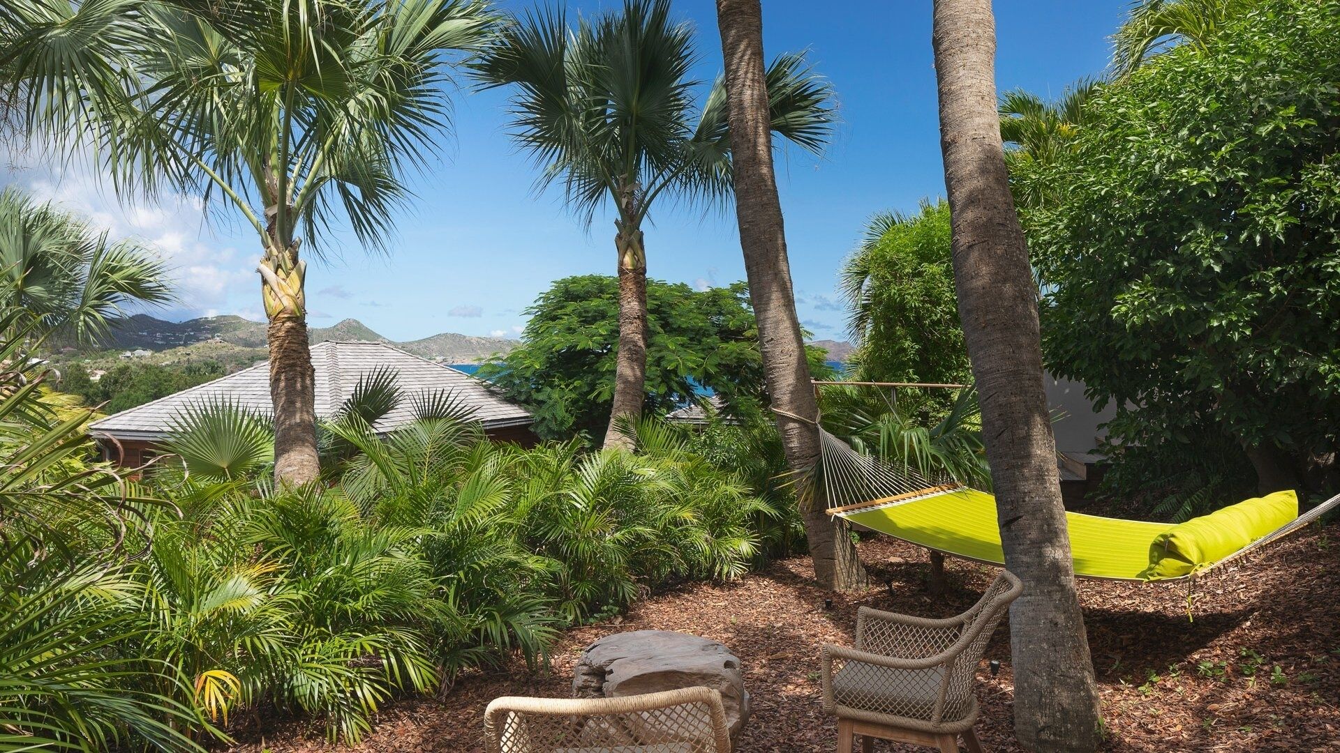 A hammock between trees, a wicker chair, and a building with a sloped roof.