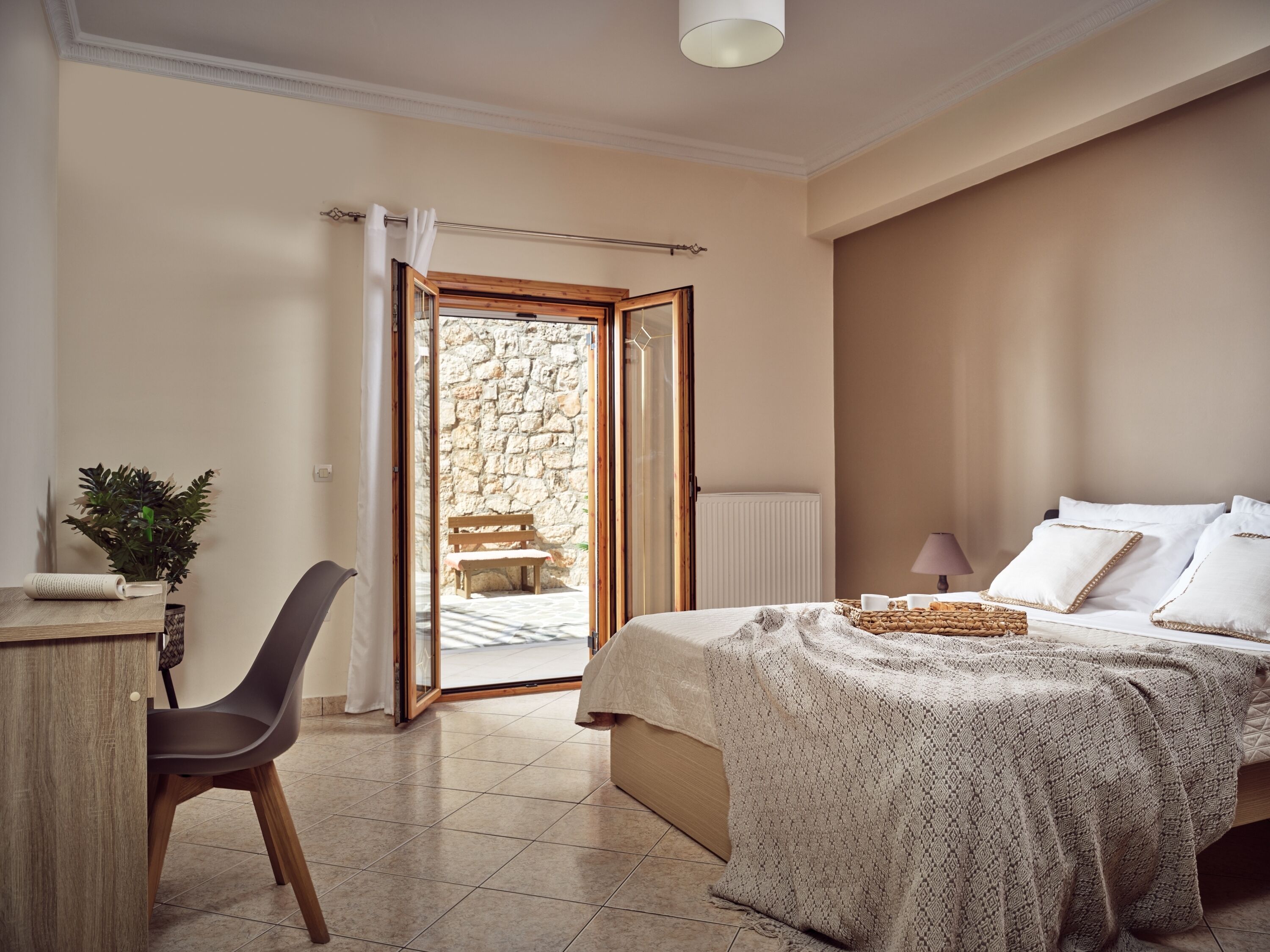 A bedroom with a bed, a desk, a chair, a radiator, and a view of an outdoor stone wall through a glass door.