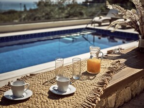Ett bord vid poolkanten med en kanna apelsinjuice, två glas och en kopp på en vävd matta.