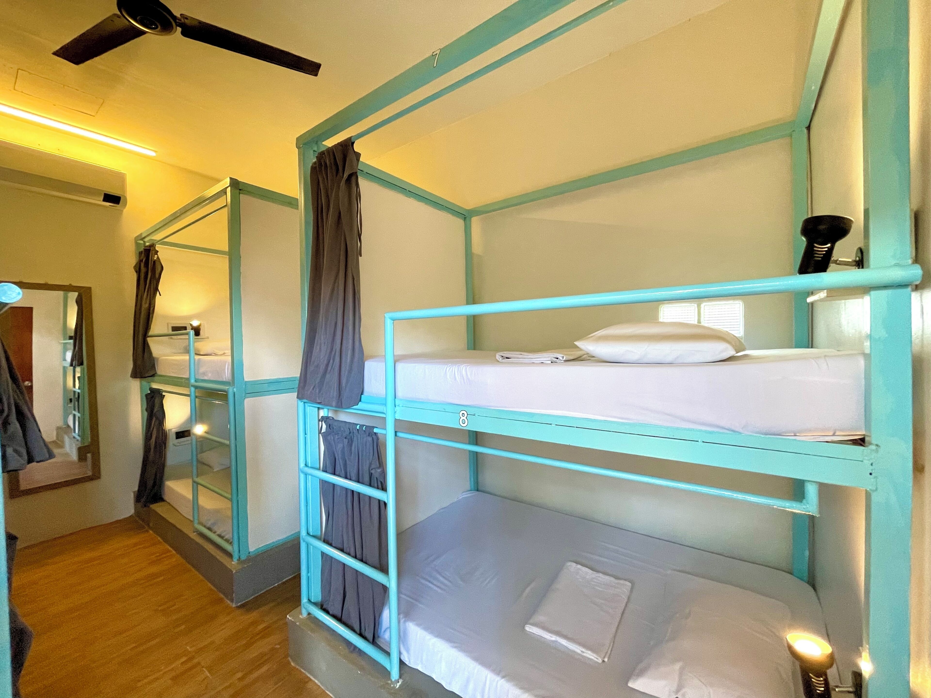 Een slaapkamer met stapelbed, plafondventilator, spiegel en raam met gordijnen.