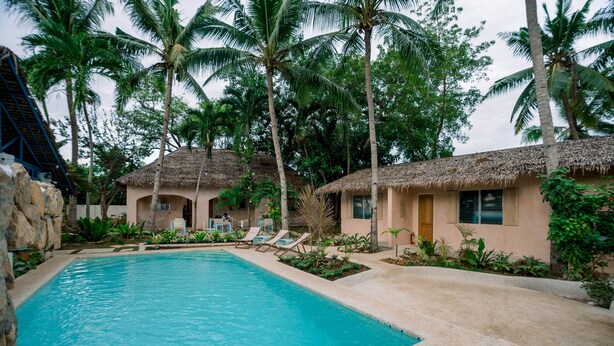 A swimming pool surrounded by thatched-roof cottages, palm trees, and a stone wall. - Mad Monkey Panglao (Dauis)
