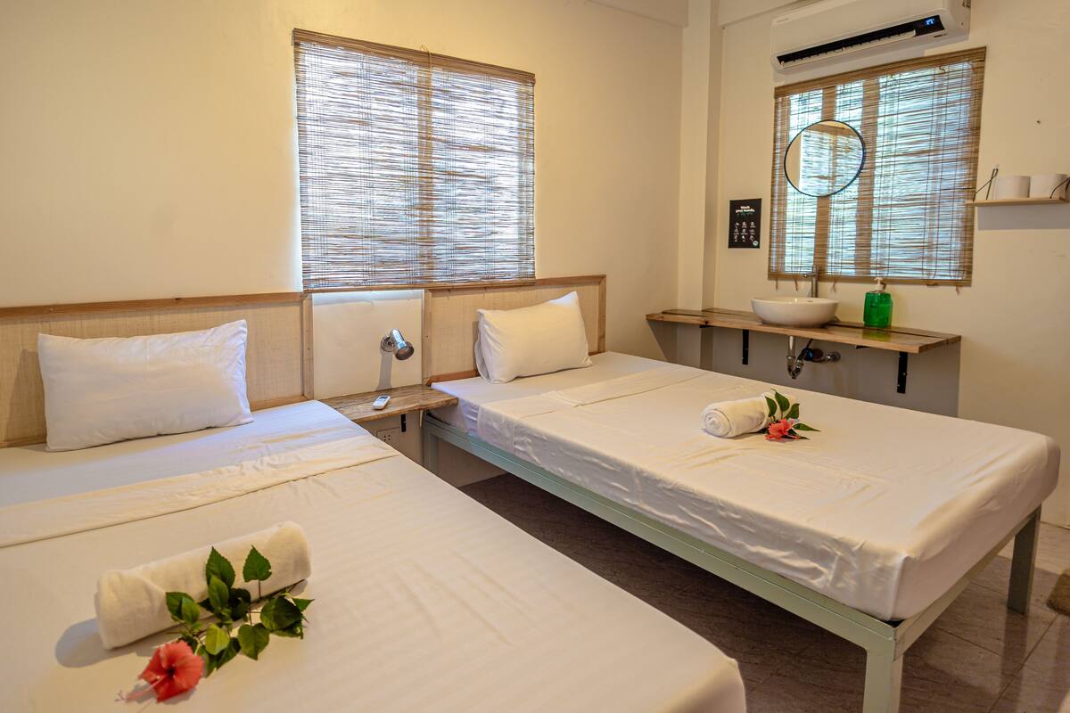 Une chambre d’hôtel avec deux lits, une table de chevet avec une lampe, une petite étagère avec des articles de toilette et une fenêtre avec des stores.