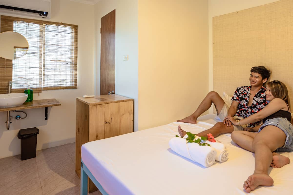 Une chambre à coucher comprenant un lit, une table de chevet, un miroir, une fenêtre avec des stores, un lavabo et une poubelle.