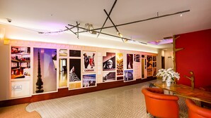 A hotel lobby with a long wall displaying various photographs, a wooden reception desk, and orange chairs.