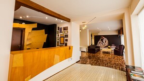 A modern hotel lobby with a wooden reception desk, a patterned carpet, and a seating area with a sofa and armchairs.