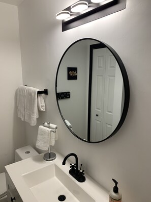 Ein Badezimmer mit einem weißen Waschbecken, einem schwarzen Wasserhahn und einem runden Spiegel. An der Wand hängen zwei weiße Handtücher und auf dem Waschbecken befindet sich ein Seifenspender.