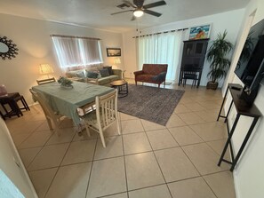 A living room with a dining area, a ceiling fan, a TV, and a large window with curtains. - Il Santuario 17 Heated Pool and Spa Close to Beach and Fifth Avenue (Naples)