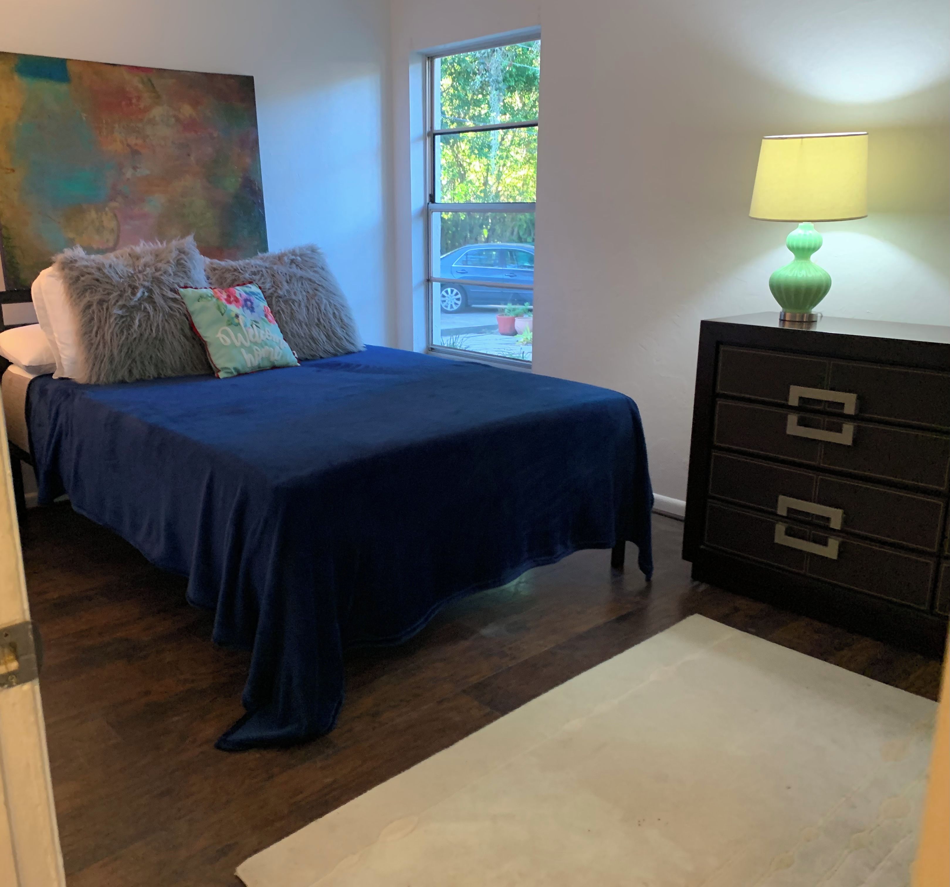 A bedroom with a bed, a dresser, a window, and a painting on the wall.