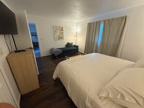 Quarto de hotel com cama, escrivaninha, cadeira, sofá, luminária e televisão.
