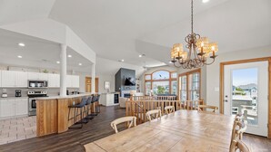 Een ruime keuken met een centraal kookeiland, een eethoek met een grote houten tafel, en uitzicht op de omgeving door grote ramen.