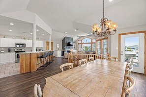 A spacious kitchen with a central island, a dining area with a large wooden table, and a view of the outdoors through large windows. - White Dolphin: Hot Tub | Rec Room | Pool Access (Corolla)