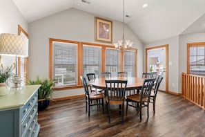 Un coin repas avec une table et des chaises en bois, de grandes fenêtres équipées de stores, un lustre et une vue sur une maison par la fenêtre.