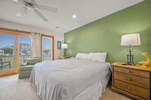 A bedroom with a bed, nightstands, a lamp, a dresser, and a view of a house through a sliding door. - White Dolphin: Hot Tub | Pool Access | Dogs OK (Corolla)
