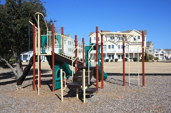 A playground with climbing structures and a slide, set against a backdrop of residential houses. - White Dolphin: Hot Tub | Pool Access | Dogs OK (Corolla)
