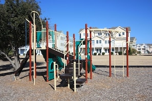 A playground with climbing structures and a slide, set against a backdrop of residential houses.