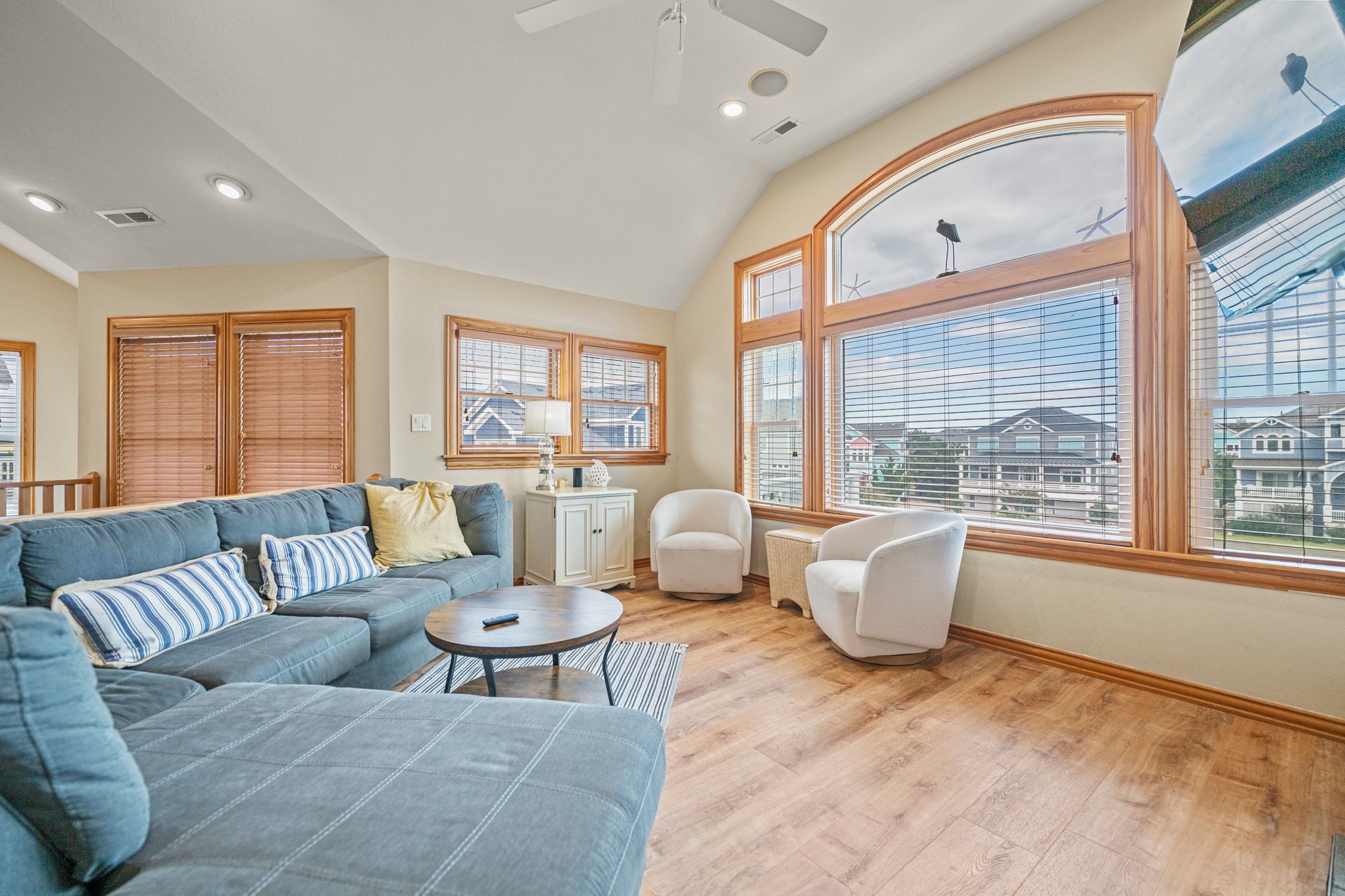 A spacious living room with a large sofa, a coffee table, and a white armchair. There are multiple windows providing views of the outside.