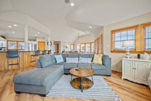 A spacious living area with a large grey sectional sofa, a round coffee table, and a kitchen area with wooden cabinets and a dining table.